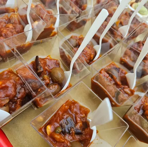 Caponata porzionata in coppette trasparenti con le forchettine di plastica, appoggiate sulla carta da forno.