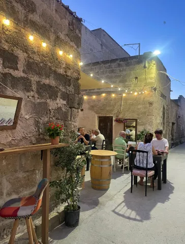 Il cortile prima che faccia buio: lampadine accese lungo il muro di tufo, un tavolo alto ricavato da una botte, gerani e piante in vaso, ospiti in piedi a chiacchierare.