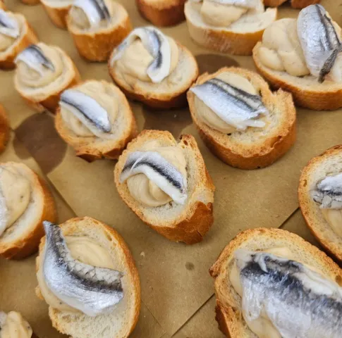 Crostini di pane allineati sulla carta da forno, ognuno con una crema chiara e sopra un filetto di pesce marinato dalla pelle argentata.