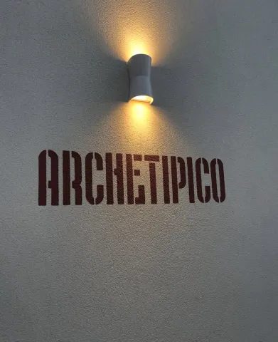 La scritta ARCHETIPICO stampata a stencil in colore ruggine sull'intonaco chiaro, illuminata dall'alto da una piccola applique che getta un cono di luce ambra.