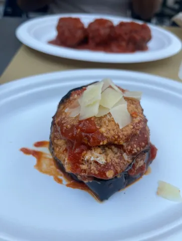 Un piatto bianco con melanzana, polpetta, salsa di pomodoro e scaglie di formaggio; sullo sfondo un secondo piatto con altre polpette al sugo.